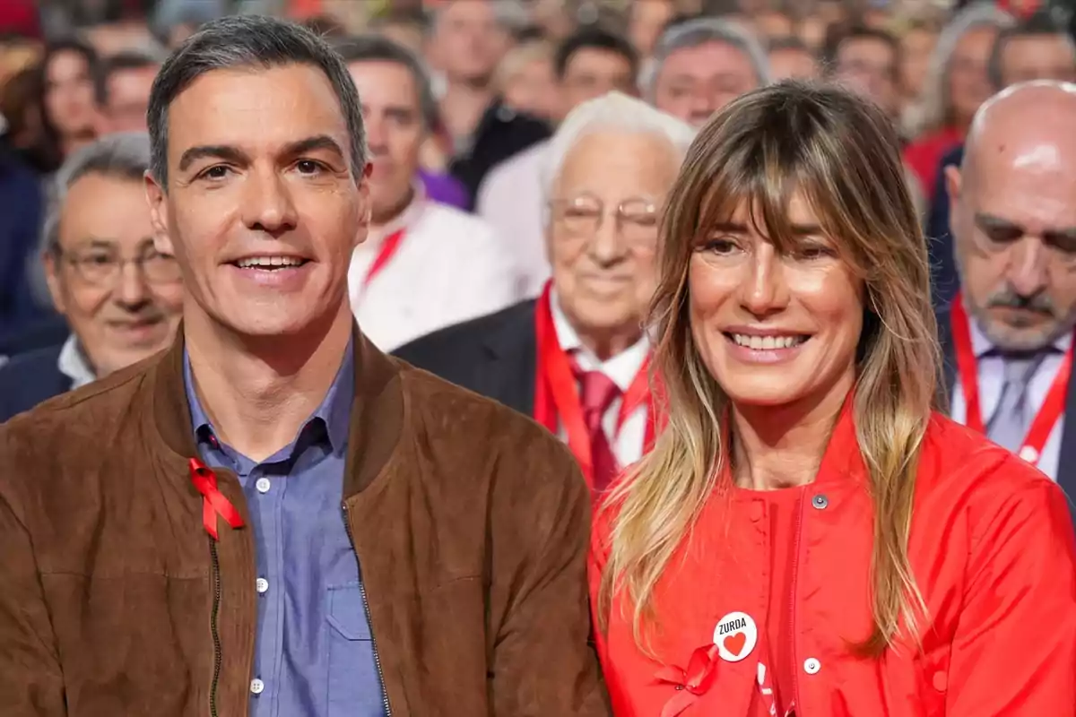 Dos personas sentadas en un evento con otras personas al fondo, ambos llevan cintas rojas.