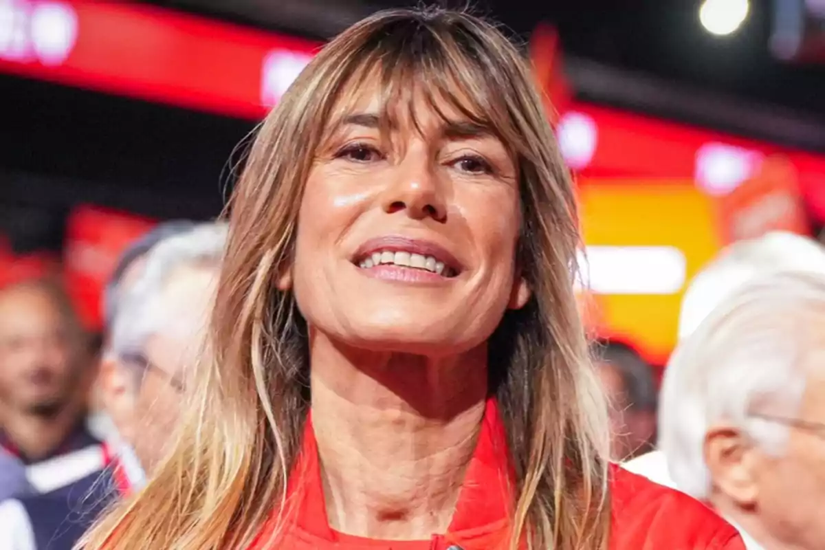 Una mujer sonriente con cabello largo y rubio en un entorno con luces rojas y personas desenfocadas al fondo.