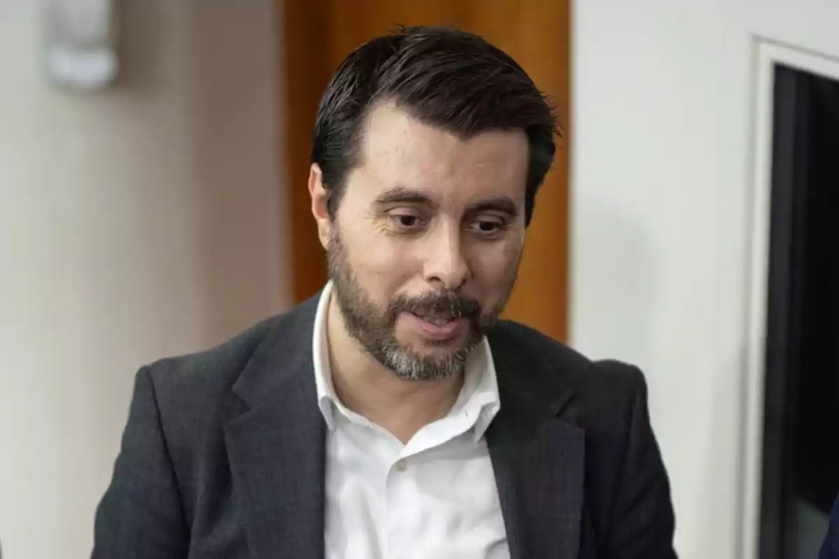 Un hombre con barba y cabello oscuro lleva un traje oscuro y camisa blanca.