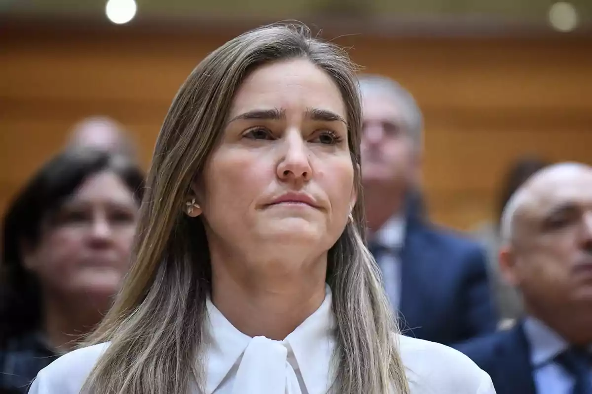 Mujer con expresión seria en un entorno formal con personas al fondo.