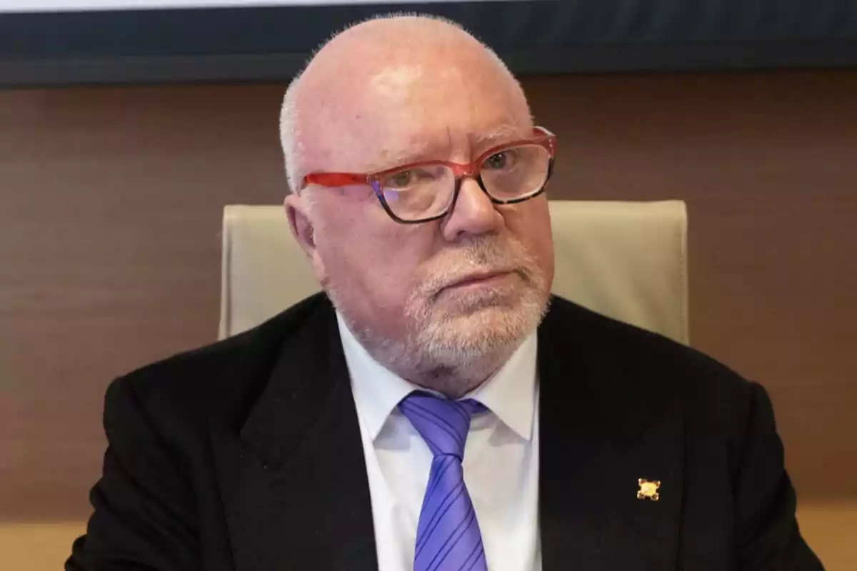Un hombre mayor con gafas y barba blanca lleva un traje oscuro y corbata morada mientras está sentado en una silla. Un hombre mayor con gafas y barba blanca lleva un traje oscuro y corbata morada mientras está sentado en una silla.