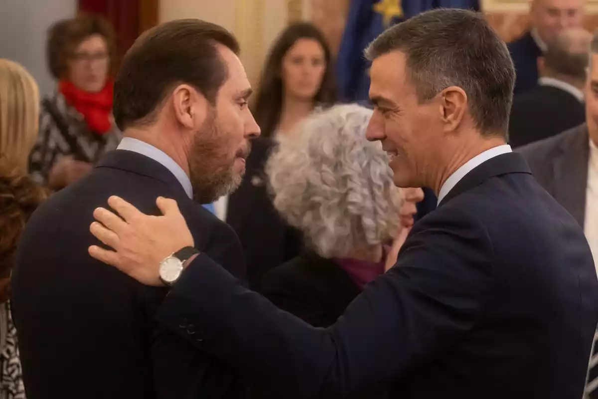 Dos hombres conversan y se saludan amistosamente en un evento formal rodeados de otras personas.