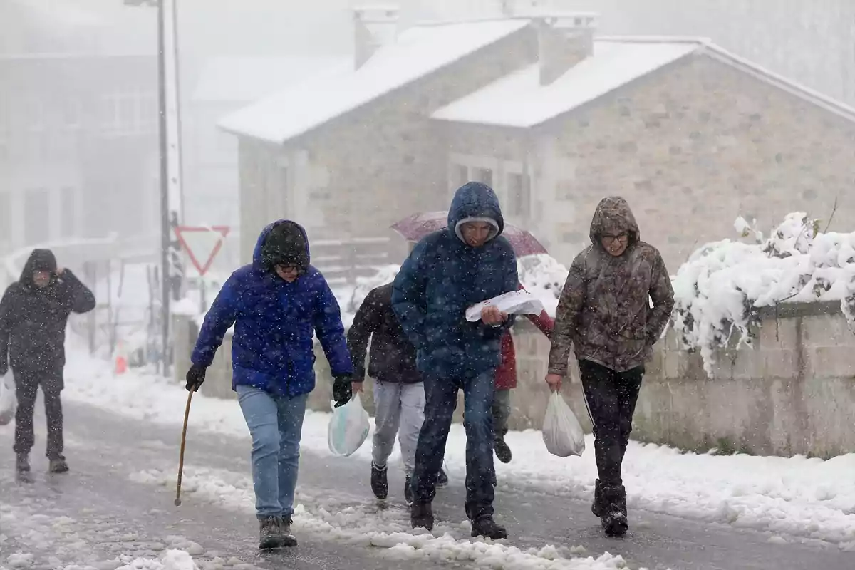 Personas caminando por una calle nevada durante una tormenta de nieve, vistiendo abrigos y capuchas para protegerse del frío.