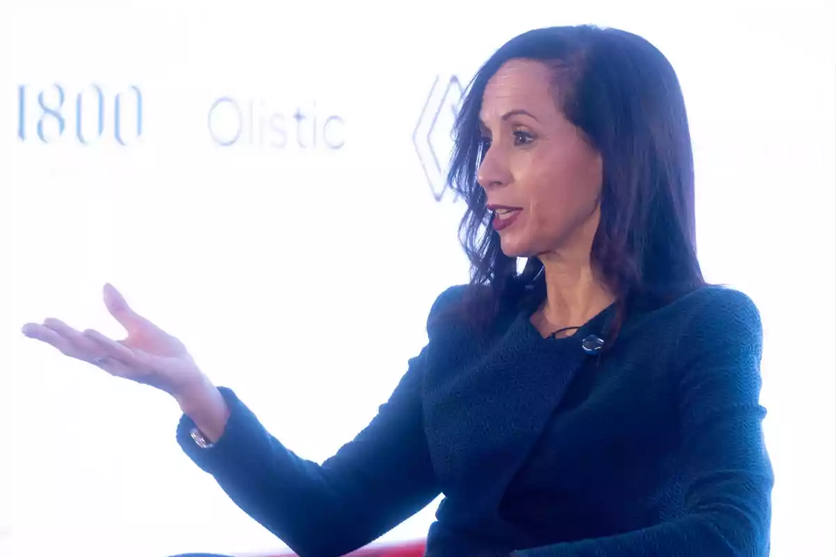 Una mujer con cabello oscuro y suelto, vestida con un suéter azul oscuro, gesticula mientras habla en un evento con un fondo blanco que muestra logotipos. Una mujer con cabello oscuro y suelto, vestida con un suéter azul oscuro, gesticula mientras habla en un evento con un fondo blanco que muestra logotipos.