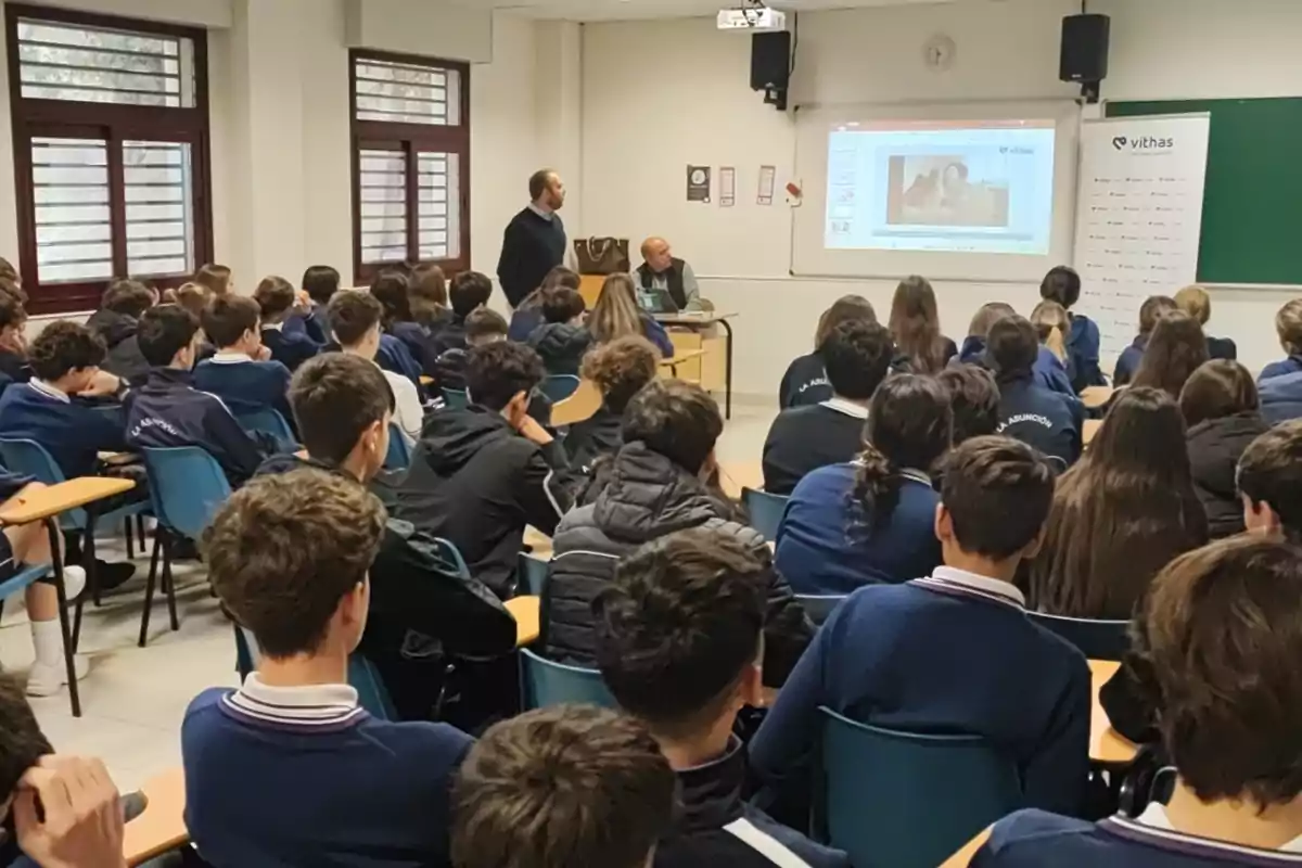 Un grupo de estudiantes en un aula presta atención a una presentación proyectada en una pantalla mientras dos personas están al frente de la clase. Un grupo de estudiantes en un aula presta atención a una presentación proyectada en una pantalla mientras dos personas están al frente de la clase.