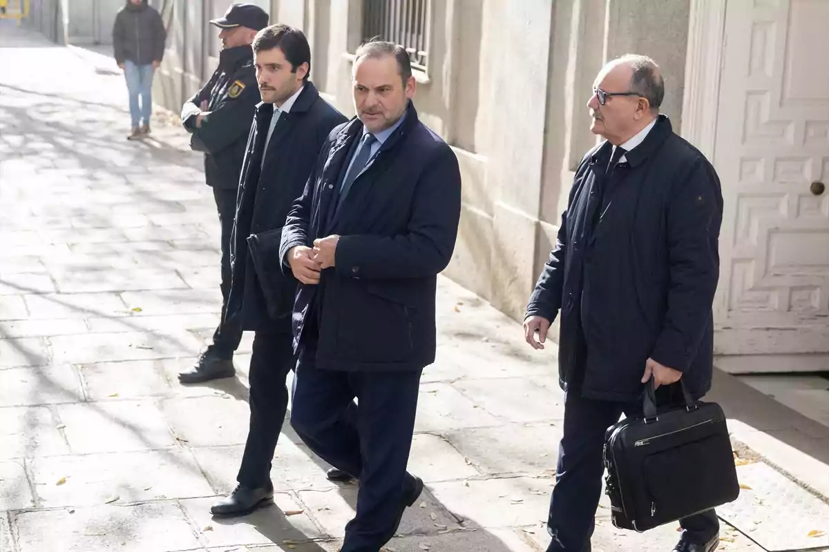 Un grupo de hombres con trajes oscuros camina por una acera junto a un edificio mientras un guardia de seguridad observa.