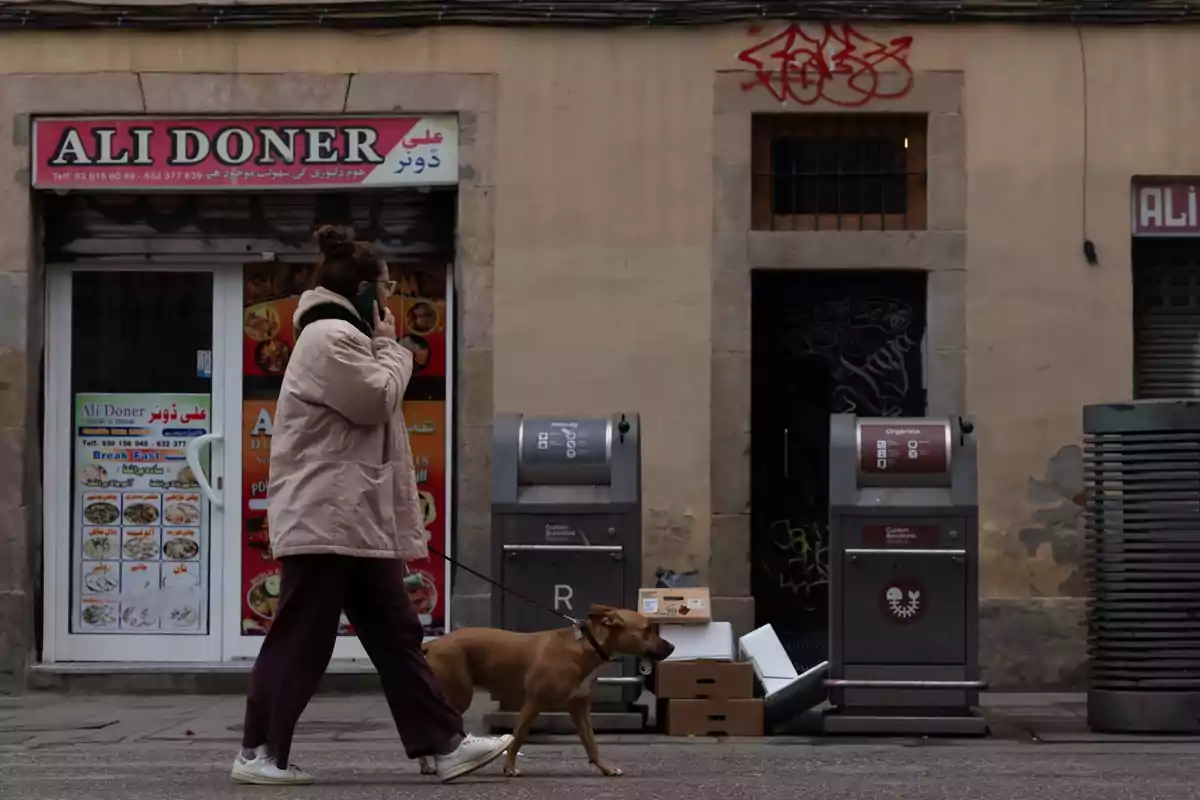 Una persona pasea a un perro frente a un restaurante llamado Ali Doner, mientras habla por teléfono y pasa junto a contenedores de basura y cajas apiladas en la acera.