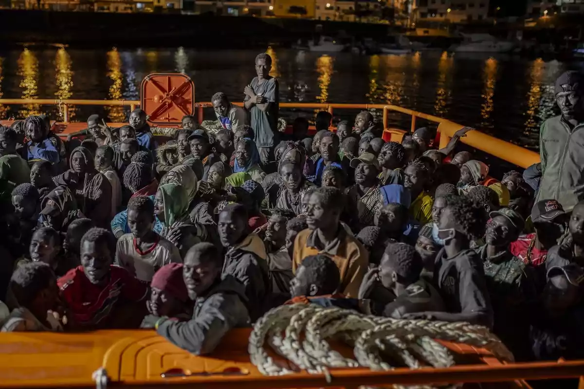 Un grupo de personas se encuentra reunido en la cubierta de un barco durante la noche con luces reflejándose en el agua al fondo.