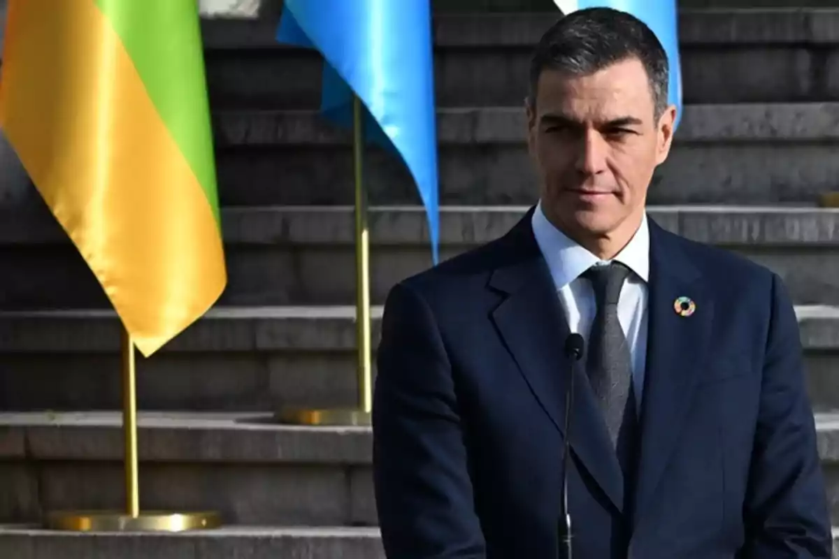Hombre de traje oscuro de pie frente a unas escaleras con banderas de colores al fondo