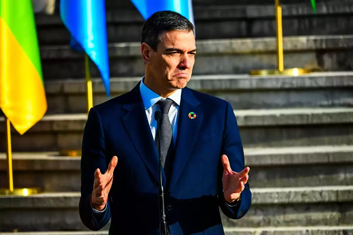 Un hombre con traje oscuro y corbata habla frente a un micrófono con banderas de fondo.