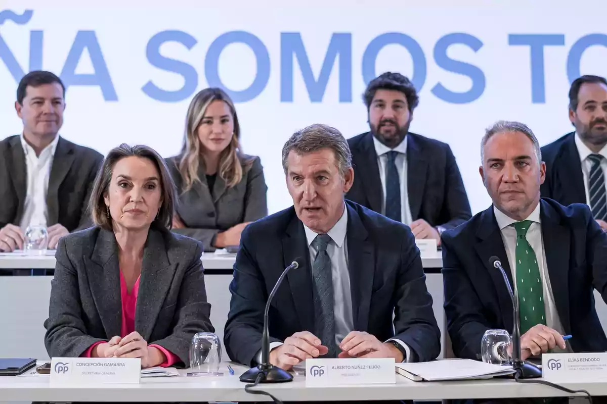 Un grupo de personas en una conferencia de prensa, sentados en una mesa con micrófonos y papeles frente a ellos, con un fondo que tiene texto parcialmente visible. Un grupo de personas en una conferencia de prensa, sentados en una mesa con micrófonos y papeles frente a ellos, con un fondo que tiene texto parcialmente visible.