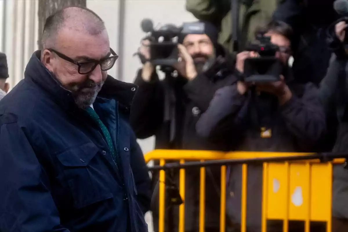 Un hombre con gafas y chaqueta oscura camina pensativo mientras varios fotógrafos y camarógrafos lo graban detrás de una valla amarilla. Un hombre con gafas y chaqueta oscura camina pensativo mientras varios fotógrafos y camarógrafos lo graban detrás de una valla amarilla.