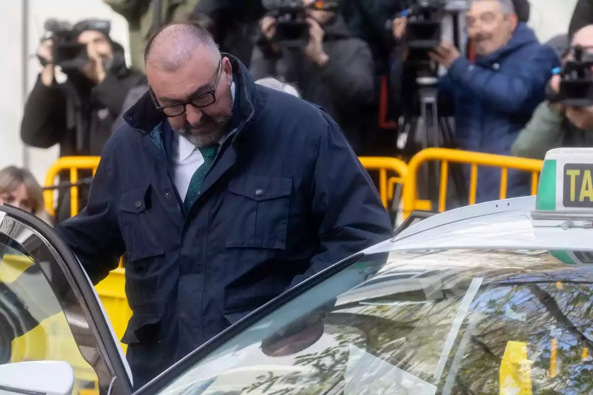 A man wearing glasses and a dark jacket approaches a taxi while several people with cameras watch him.