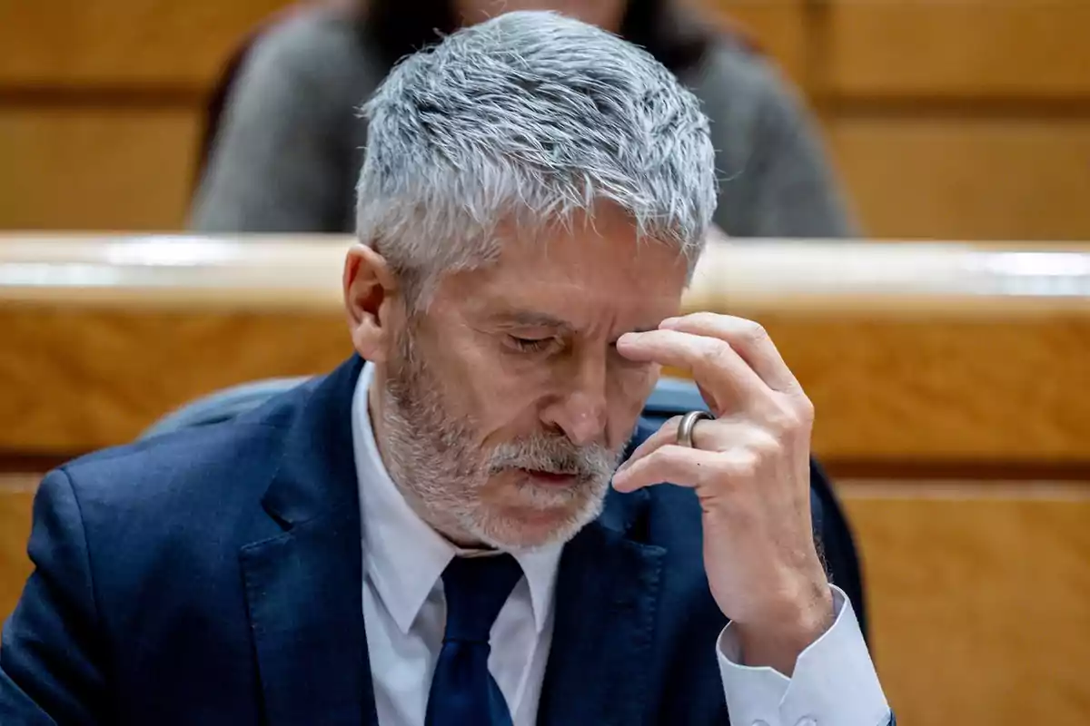 Un hombre de cabello canoso y barba vestido con traje azul oscuro y corbata se toca la frente con expresión pensativa en un entorno formal.