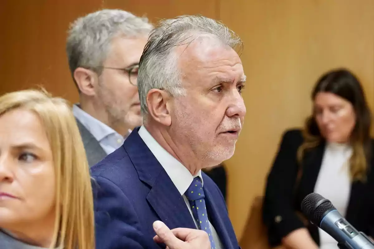 Un hombre de traje azul y corbata habla frente a un micrófono mientras otras personas están en el fondo. Un hombre de traje azul y corbata habla frente a un micrófono mientras otras personas están en el fondo.