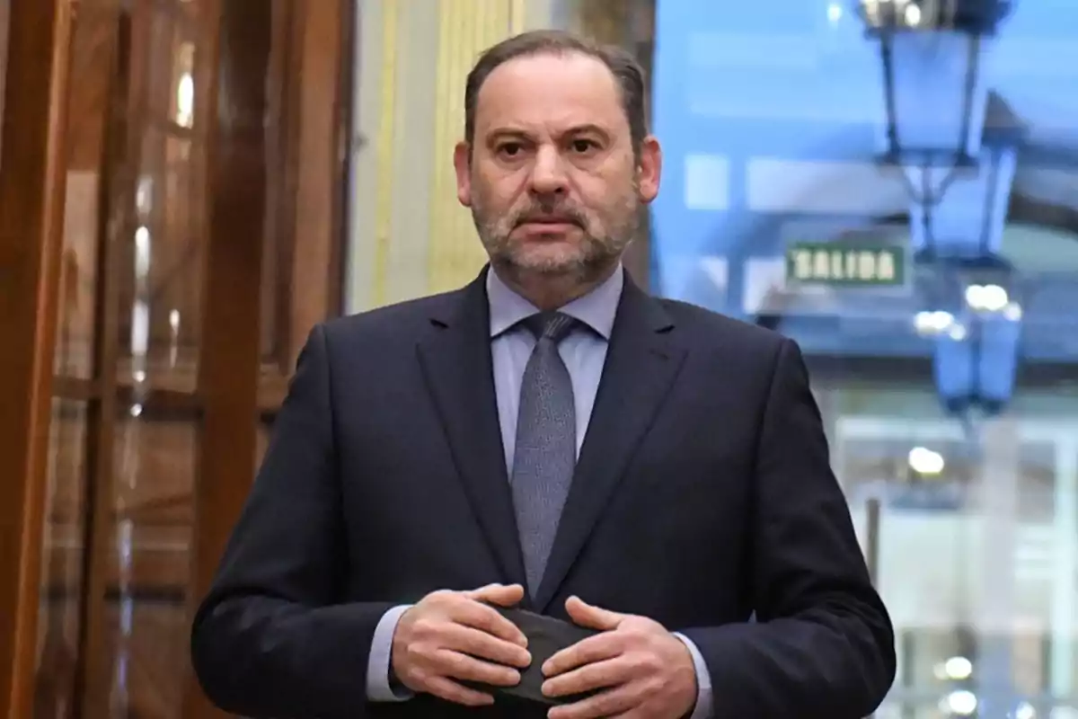 Un hombre de traje oscuro y corbata gris está de pie en un interior con una señal de salida visible al fondo.