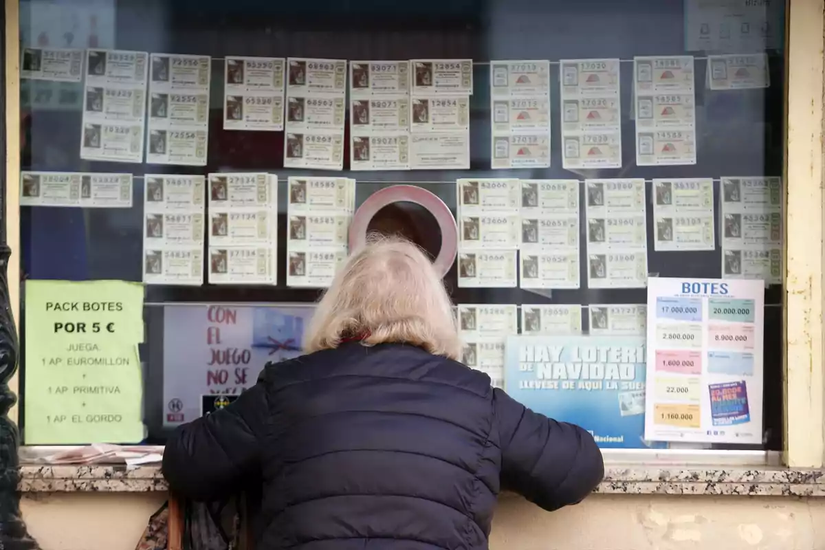 Una persona de espaldas comprando boletos de lotería en una ventanilla con varios carteles y números expuestos.