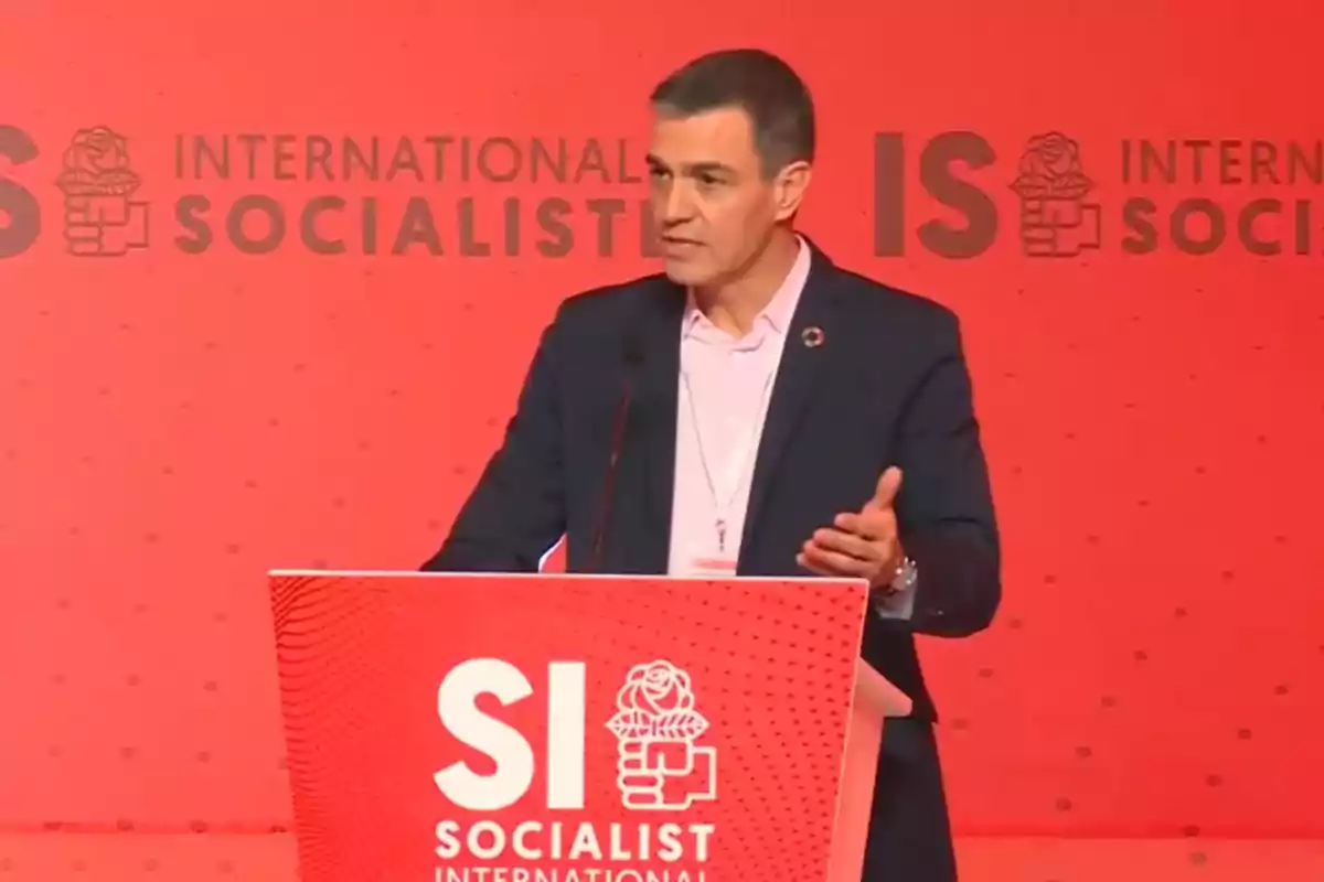 Un hombre hablando en un podio con el logotipo de la Internacional Socialista sobre un fondo rojo.