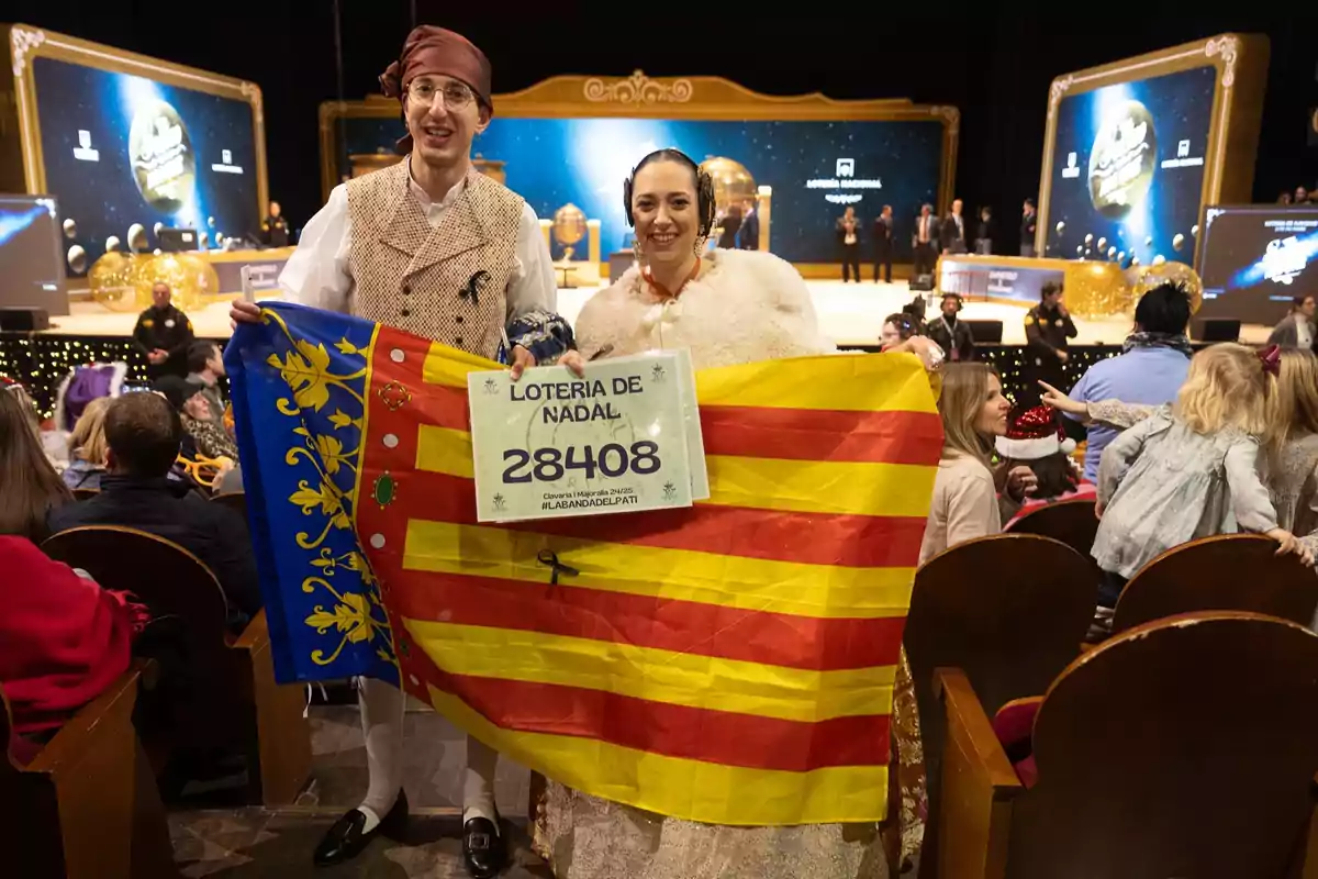 Dos personas vestidas con trajes tradicionales sostienen una bandera y un cartel con el número 28408 en un evento de la Lotería de Navidad, rodeados de un público atento en un auditorio decorado festivamente.