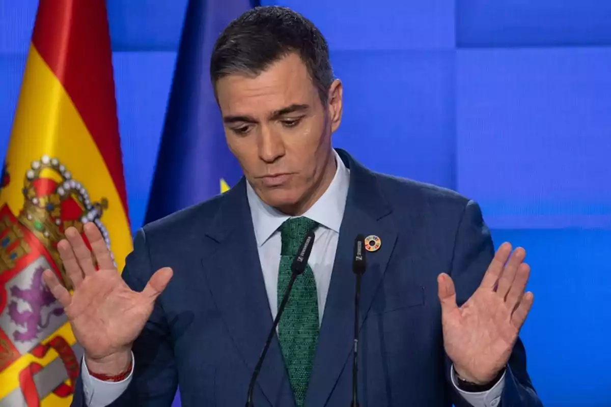 Un hombre en traje azul con corbata verde gesticula con las manos frente a dos micrófonos, con las banderas de España y la Unión Europea de fondo.