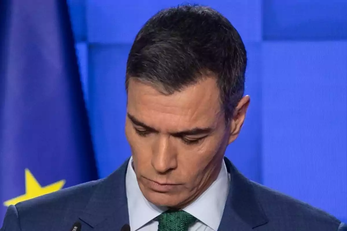 Un hombre con traje azul y corbata verde mira hacia abajo frente a un fondo azul con una bandera de la Unión Europea.