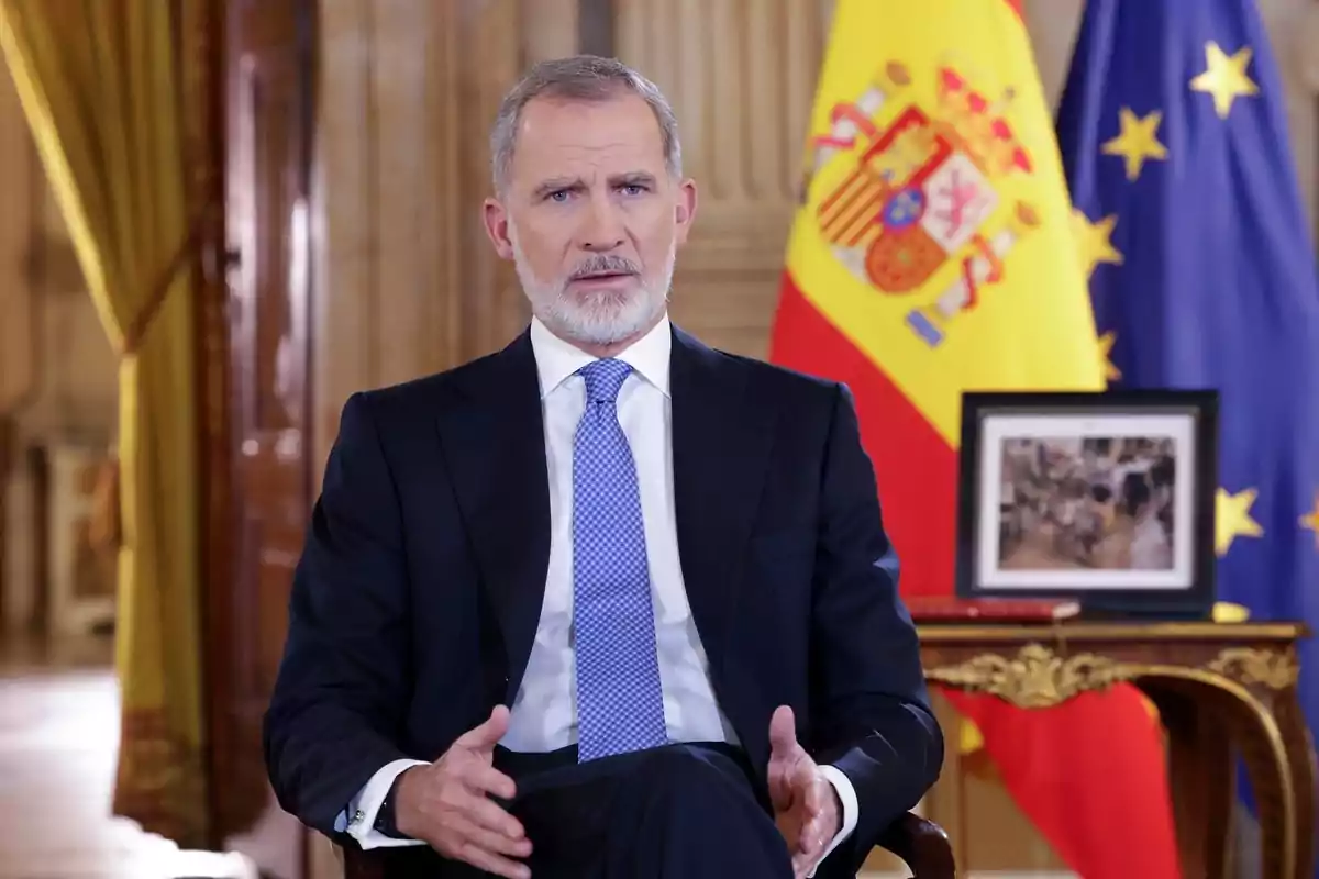 Un hombre con traje y corbata azul está sentado frente a las banderas de España y la Unión Europea en una sala elegante.