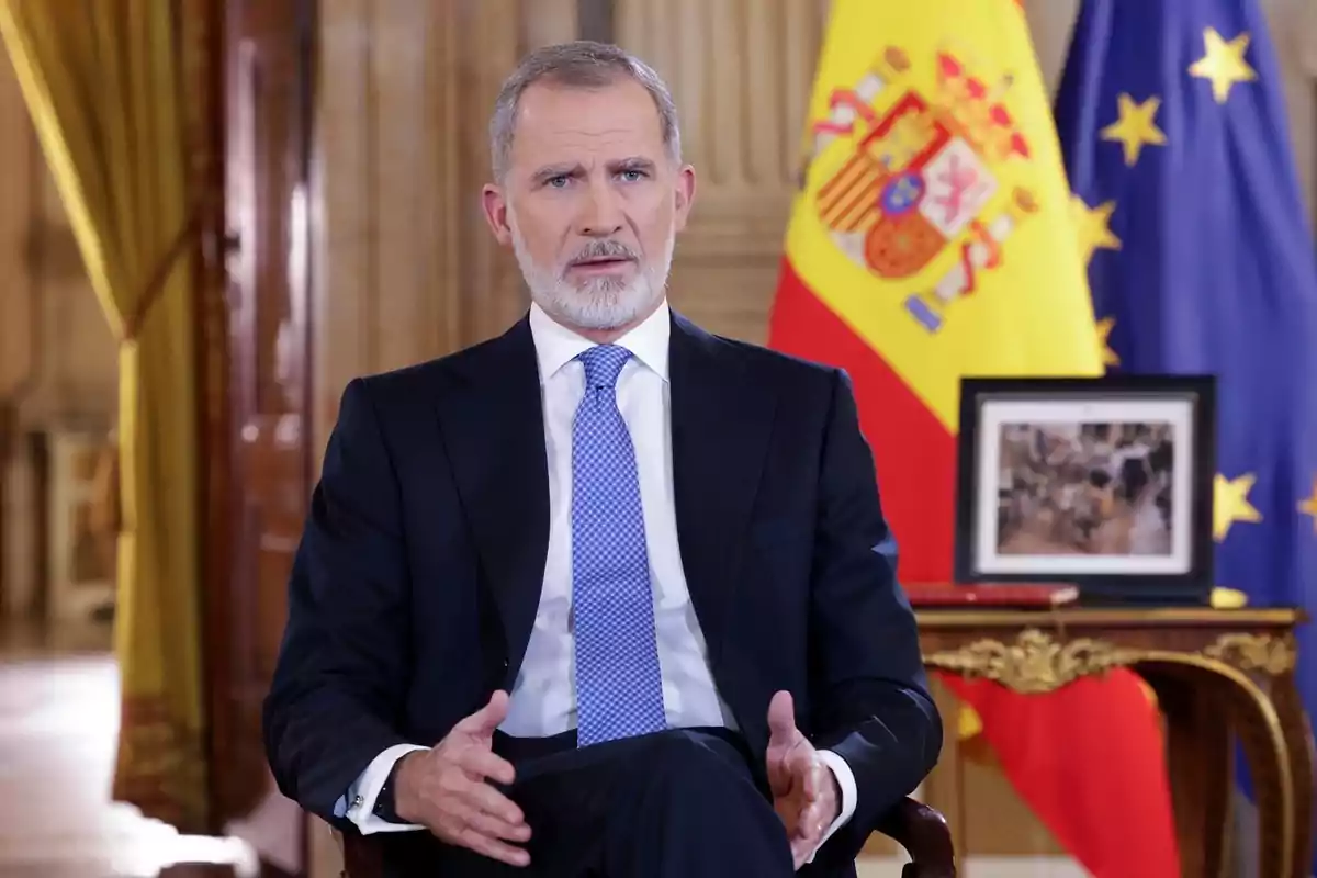 Un hombre con traje y corbata está sentado frente a las banderas de España y la Unión Europea en una sala elegante.