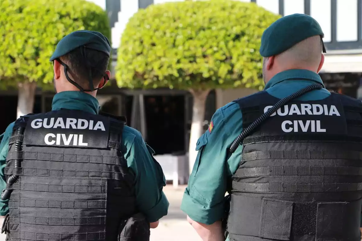 Dos agentes de la Guardia Civil de espaldas con uniformes y chalecos antibalas, en un entorno exterior con árboles.