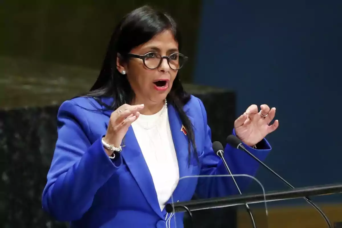 A person with glasses and a blue suit speaking at a podium with microphones.