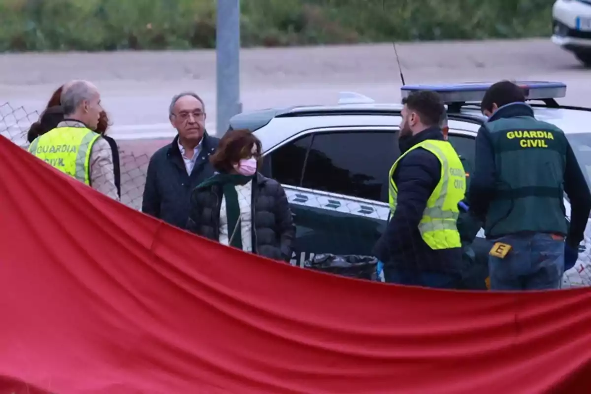 Un grupo de personas, incluyendo miembros de la Guardia Civil, se encuentra reunido junto a un vehículo oficial detrás de una barrera roja.