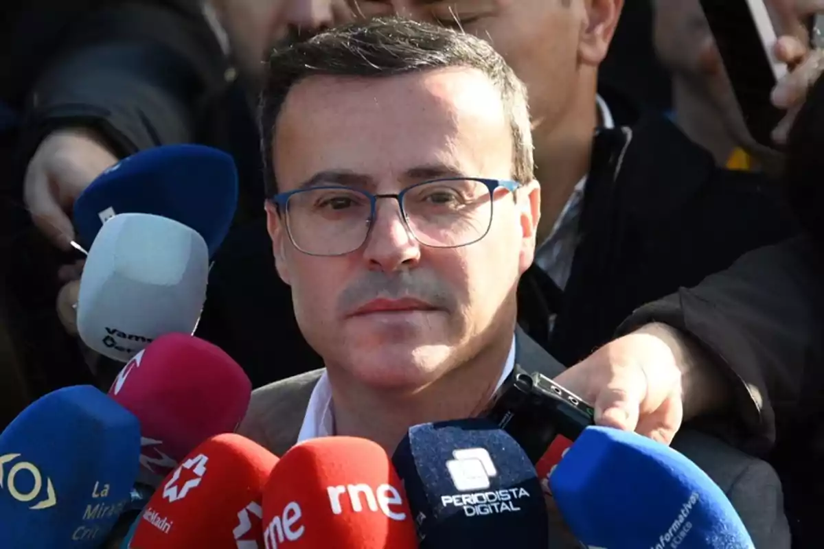 Un hombre con gafas rodeado de micrófonos de diferentes medios de comunicación.