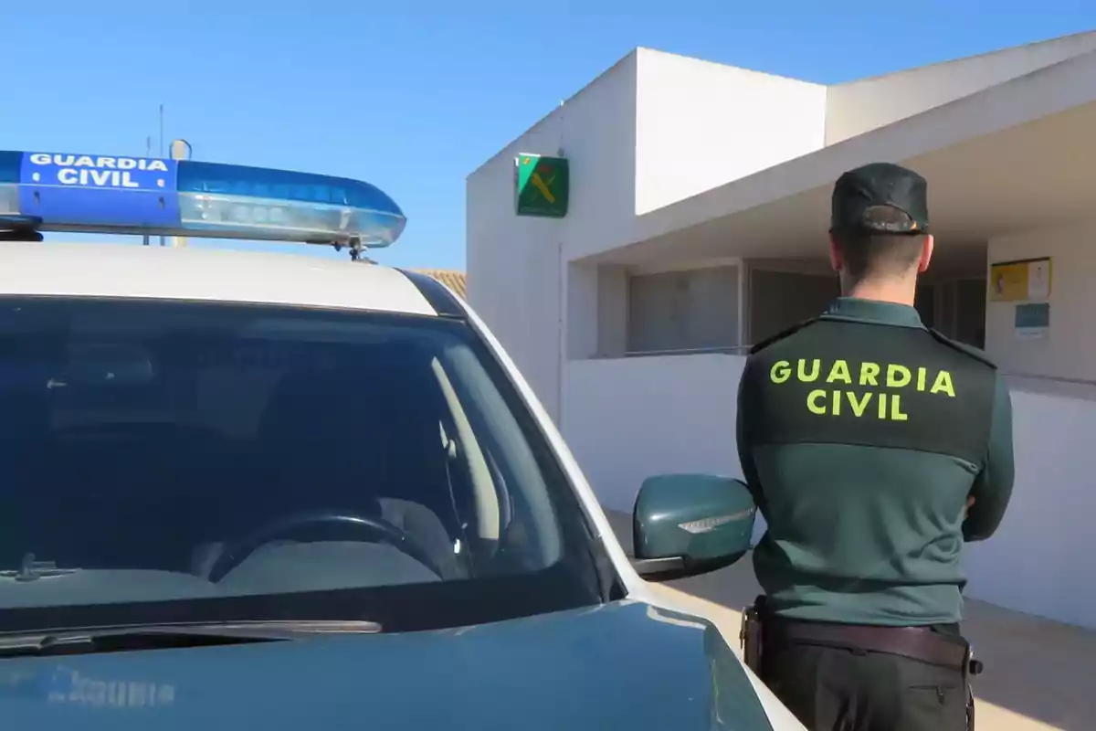 Un agente de la Guardia Civil de espaldas junto a un vehículo oficial estacionado frente a un edificio.