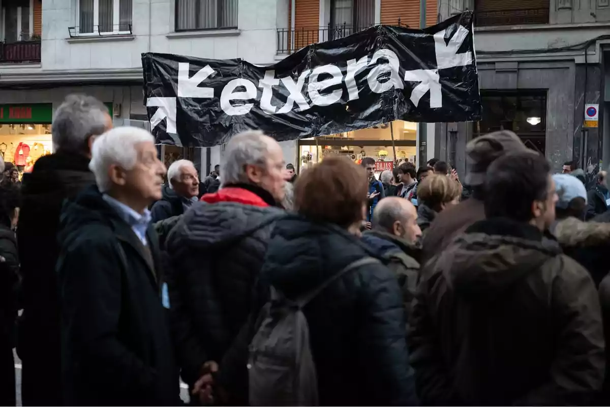 A group of people gather on a street in front of a black sign with the word A group of people gather on a street in front of a black sign with the word