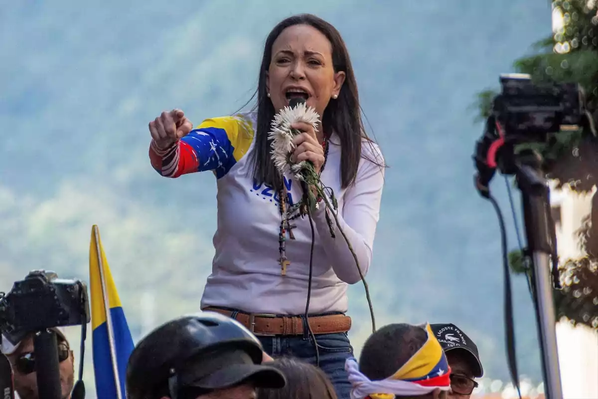 Una persona hablando en un evento al aire libre, sosteniendo flores y usando una camiseta con los colores de la bandera de Venezuela. Una persona hablando en un evento al aire libre, sosteniendo flores y usando una camiseta con los colores de la bandera de Venezuela.