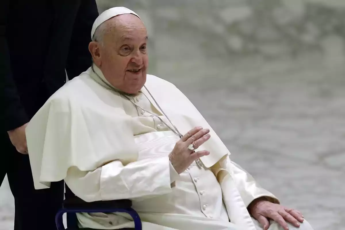 An elderly man dressed in white religious attire is sitting in a wheelchair, raising one hand while smiling.