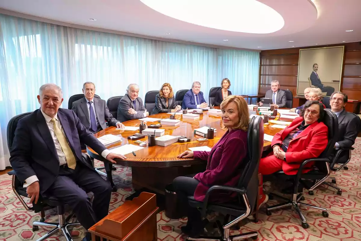 Un grupo de personas sentadas alrededor de una mesa de conferencias en una sala de reuniones con cortinas y un retrato en la pared. Un grupo de personas sentadas alrededor de una mesa de conferencias en una sala de reuniones con cortinas y un retrato en la pared.