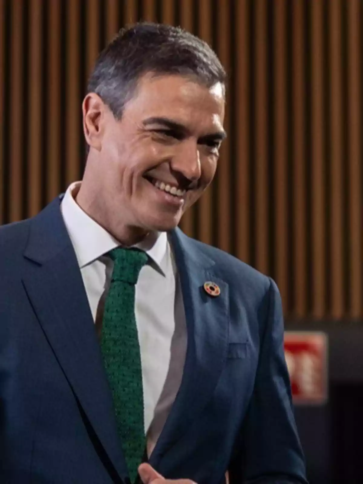 A man in a blue suit and green tie smiles in an interior setting with a wooden background.