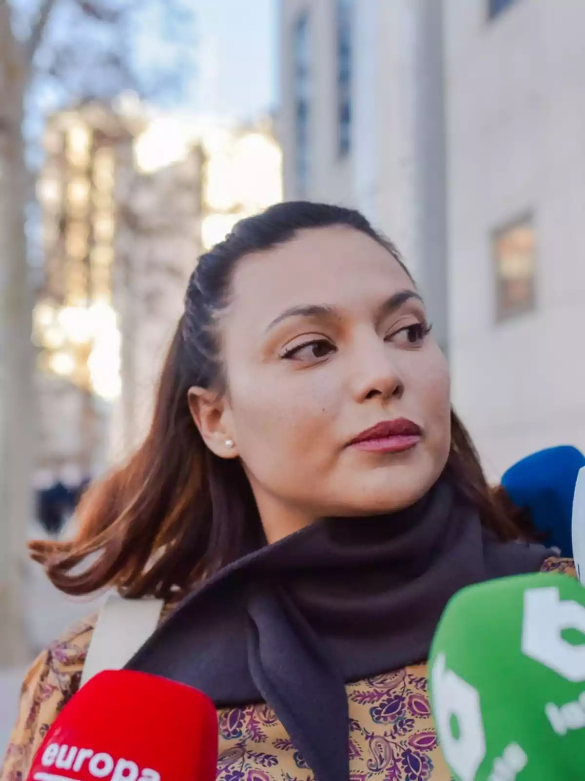 Woman with dark scarf surrounded by different colored microphones in an urban setting.