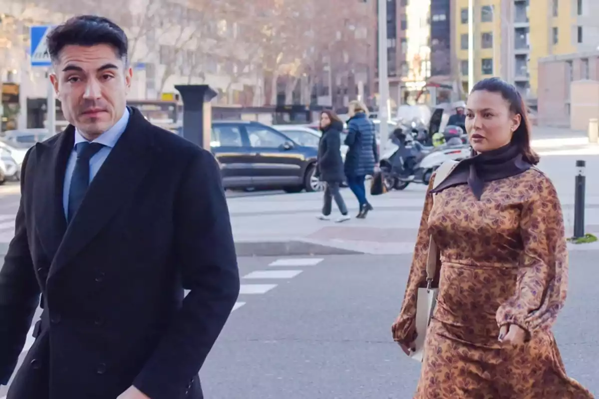 Two people walking down an urban street with buildings and vehicles in the background.