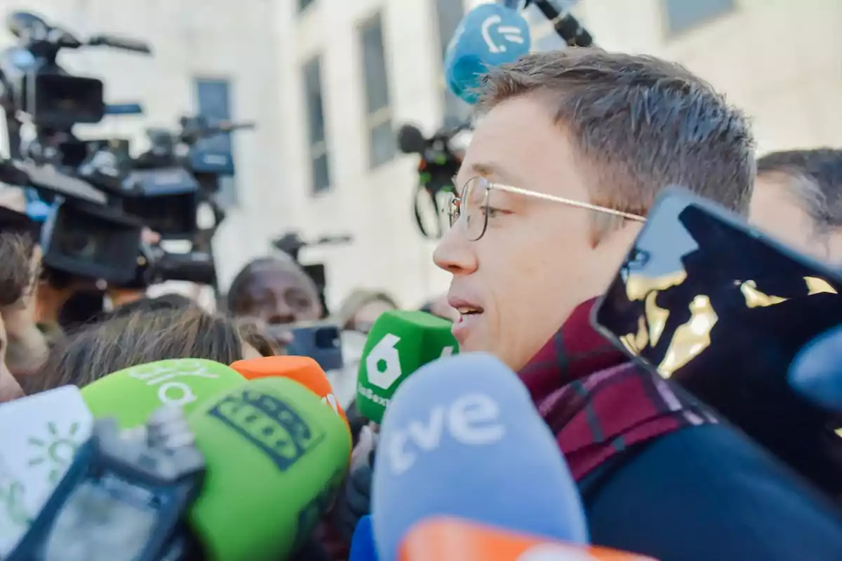 A man with glasses surrounded by microphones from different media outlets and cameras in an urban setting.