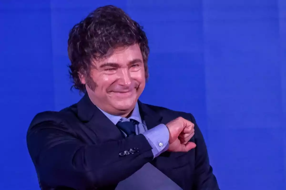 A man in a dark suit and blue jersey smiles while gesturing with his arm in front of a blue background.