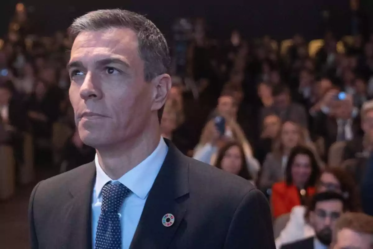 A man in a dark suit and blue tie at an event with an audience in the background. A man in a dark suit and blue tie at an event with an audience in the background.