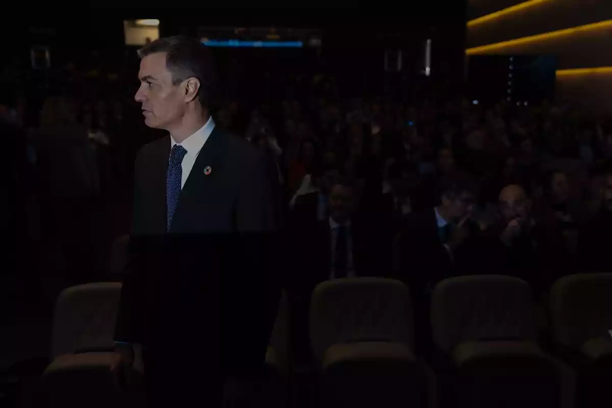 A man in a dark suit and blue tie stands in a crowded auditorium with dim lights and a formal atmosphere.