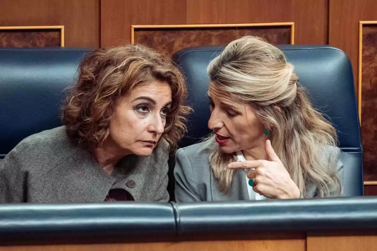 Dos mujeres sentadas en un entorno formal conversando de manera seria. Dos mujeres sentadas en un entorno formal conversando de manera seria.