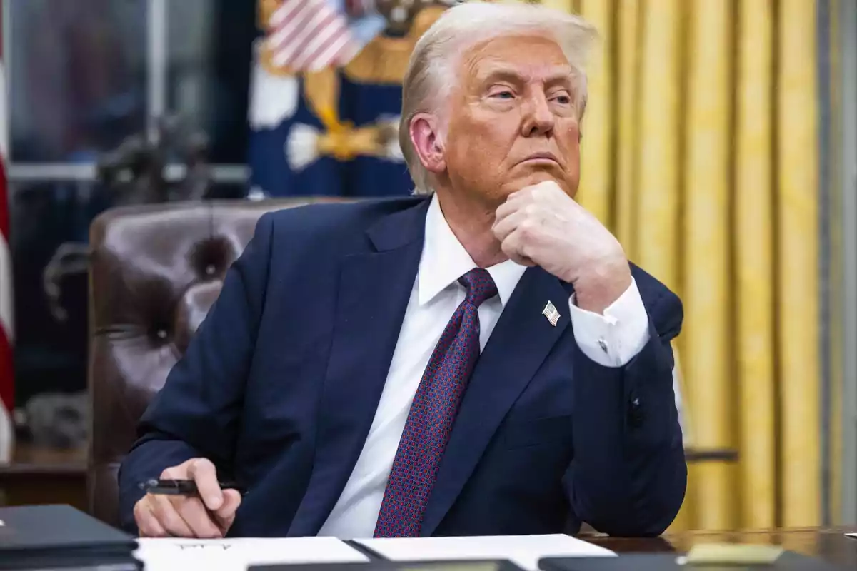 A man in a dark suit and red tie sits at a desk with a thoughtful expression, holding a pen in one hand.