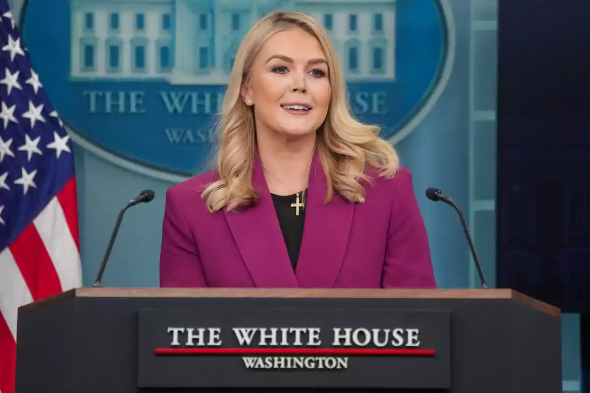 A blonde-haired woman in a purple jacket speaks from a podium at the White House.