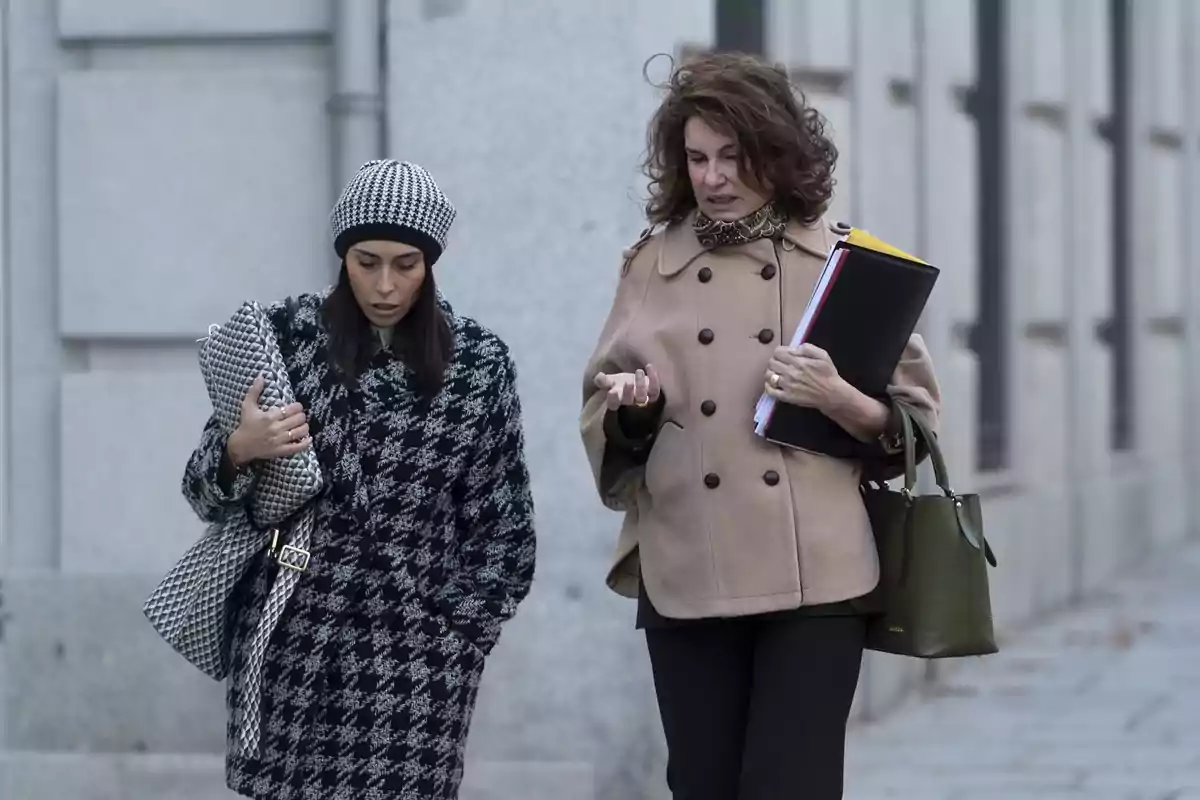 Dos mujeres caminando por la calle, una lleva un abrigo y gorro de cuadros y la otra un abrigo beige, mientras conversan y llevan documentos. Dos mujeres caminando por la calle, una lleva un abrigo y gorro de cuadros y la otra un abrigo beige, mientras conversan y llevan documentos.