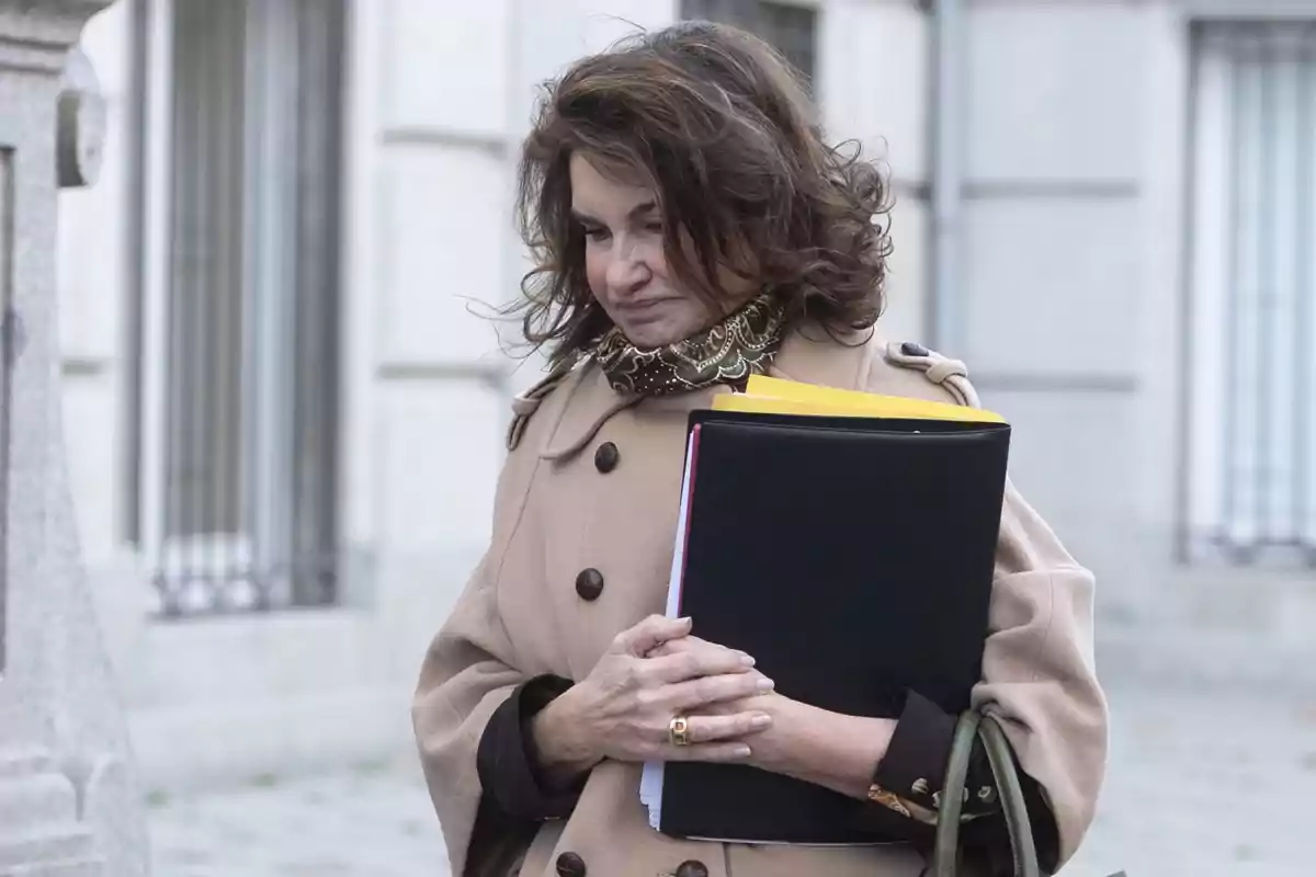 Mujer con abrigo beige y bufanda estampada sostiene carpetas mientras camina por la calle.