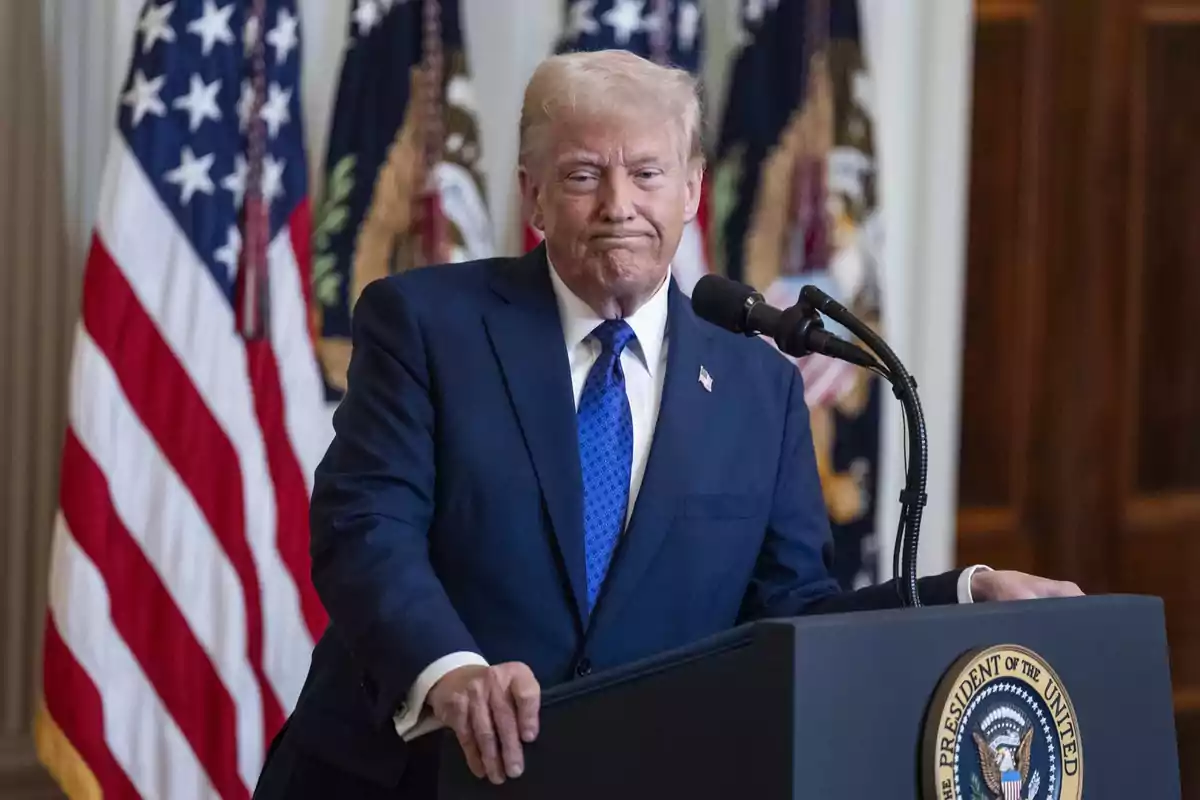 Un hombre de traje azul oscuro y corbata azul está de pie frente a un podio con el sello presidencial, con banderas estadounidenses de fondo.