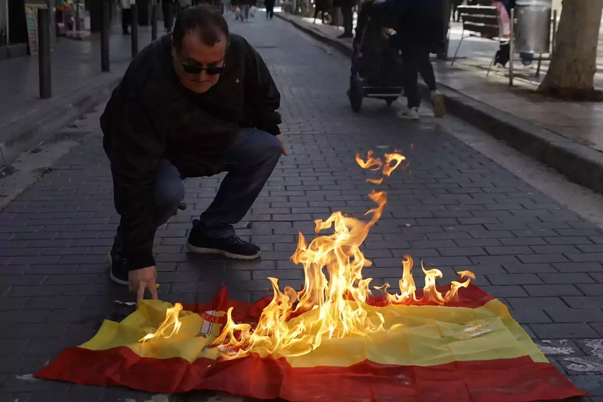 A man with sunglasses crouches in a street while a flag on the ground is on fire.